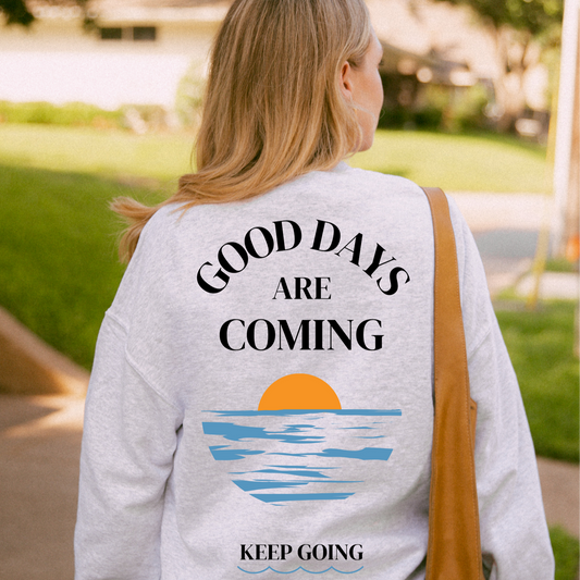 Person wearing a sweatshirt with 'Good days are coming, keep going' text and sunset design.