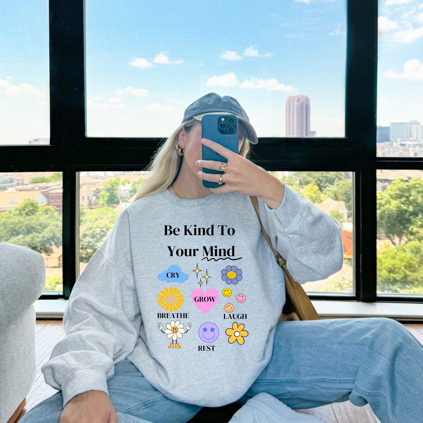 Person wearing a sweatshirt with motivational text and emojis, sitting by a window with a cityscape view.
