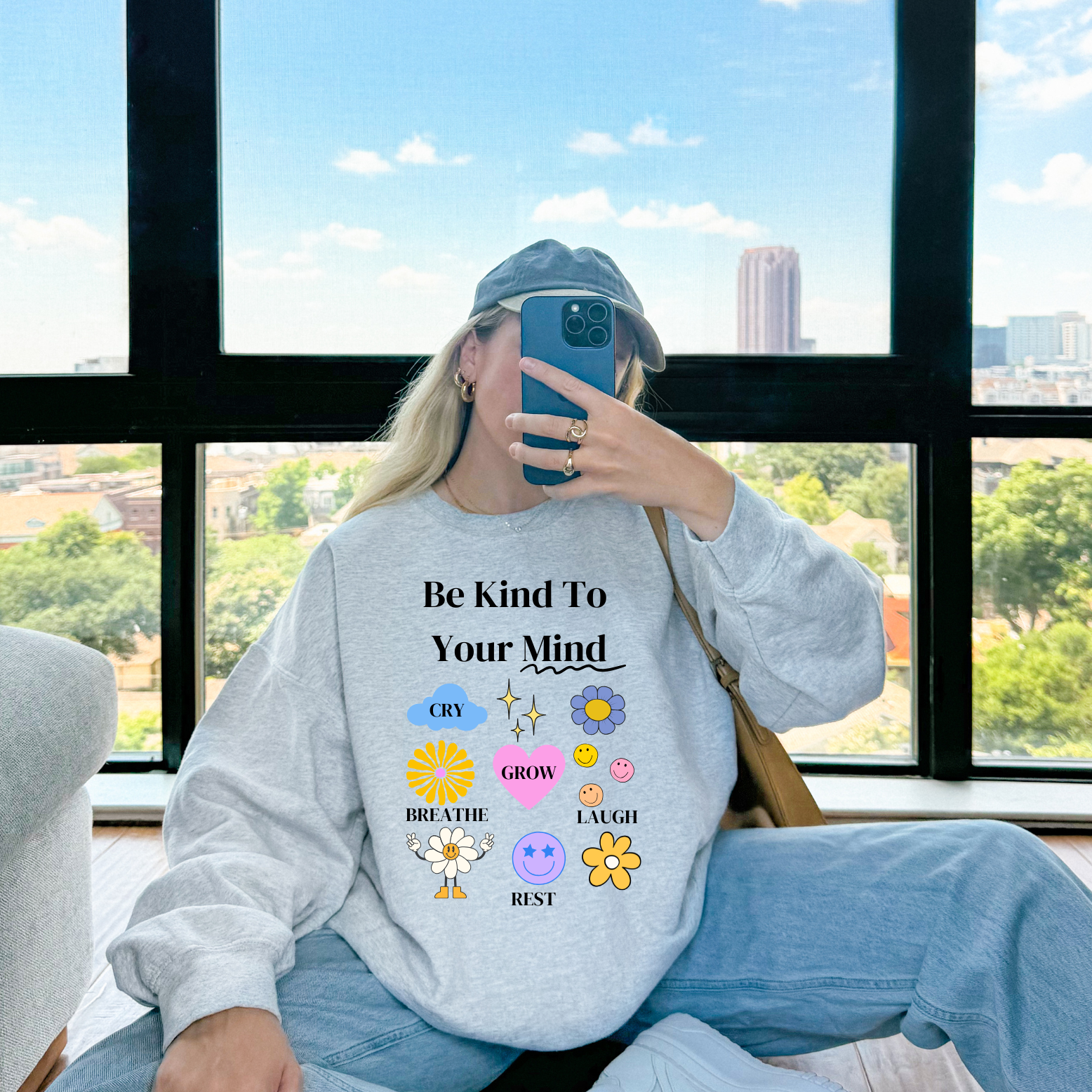Person wearing a sweatshirt with motivational text and emojis, sitting by a window with a cityscape view.